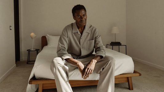 A man in his early sixties sits thoughtfully at the edge of his bed in the early morning
