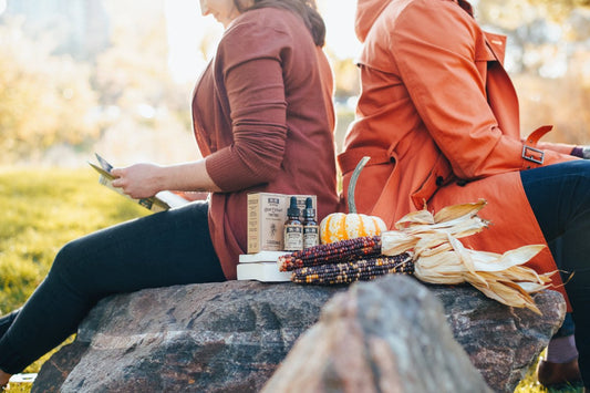 Two women sitting back-to-back outdoors with natural remedies for bacterial vaginosis including supplements, oils, and herbs placed on a rock beside them