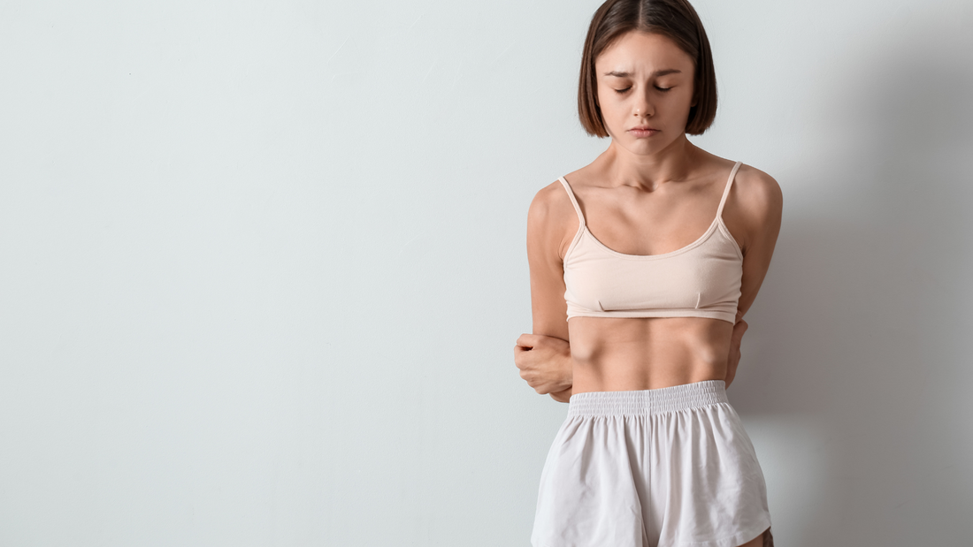 thin young woman with anorexia standing against white wall looking down