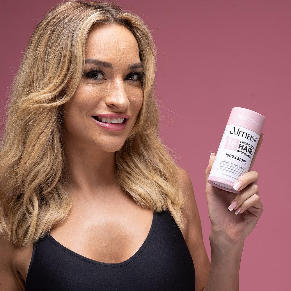 woman holding Almasi Probiotics Skin Hair & Nails supplement bottle against pink background