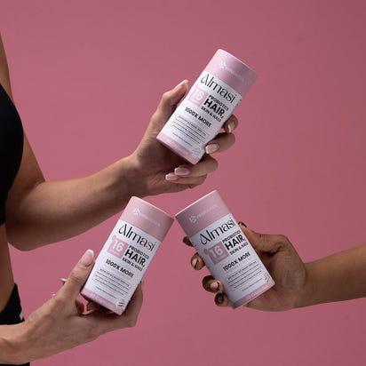 Hands holding Almasi Probiotics Skin Hair & Nails supplement bottles with pink labels against pink background